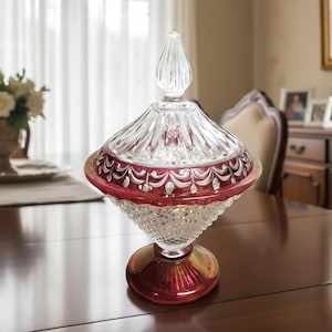 May include: A decorative glass candy dish with a lid. The dish has a clear, faceted base and a red-tinted, scalloped rim. The lid is clear with a pointed finial. The dish is sitting on a wooden table.