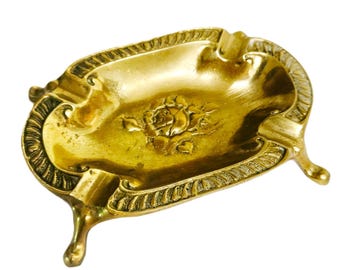 Pode incluir: Um cinzeiro oval de latão com um design central de rosa em relevo e uma borda decorativa em estilo de corda. A peça é apoiada por quatro pequenas pernas curvas e inclui entalhes integrados ao longo da borda para apoiar objetos.