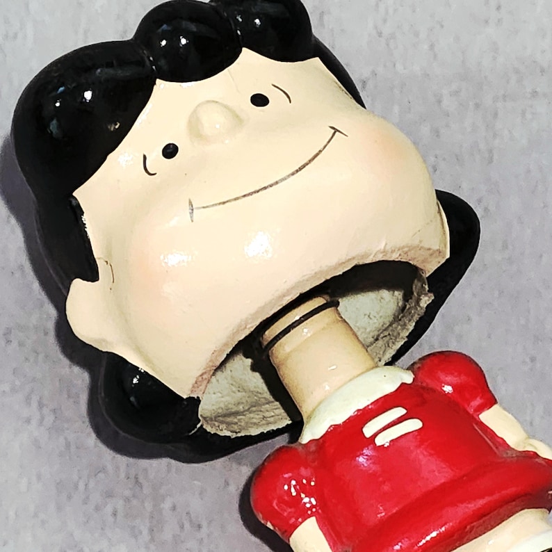 May include: A ceramic bottle stopper featuring a cartoon character's head with black hair, a smiling face, and a red dress. The stopper is designed to fit into a bottle opening.