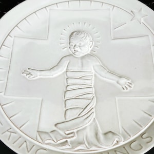 May include: White ceramic plate with a raised relief design. The design features a baby with a halo, arms outstretched, in front of a cross. A star is visible in the upper right. The words "KING OF KINGS" are along the bottom edge.