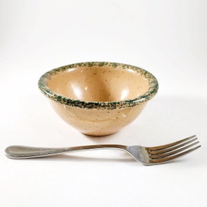 May include: A small, beige ceramic bowl with a green speckled rim. The bowl is sitting on a white surface next to a silver fork.