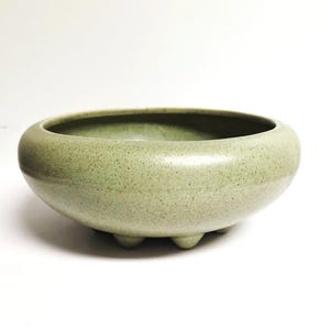 May include: A round, low-profile ceramic bowl with a speckled light green glaze. The vessel features a smooth, curved rim and is supported by three small, rounded feet at the base, providing a stable foundation for this functional piece.