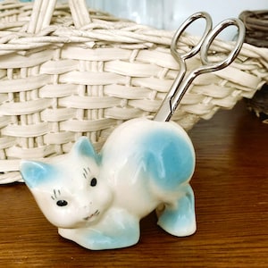 May include: A ceramic cat-shaped scissors holder in white and light blue. The cat is in a playful pose, with a pair of silver scissors inserted in its back. The holder is on a wooden surface, with a woven basket in the background.