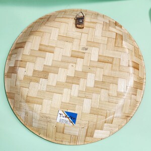 May include: A round, woven bamboo plate with a natural light brown color. The plate has a basket weave pattern and a small metal hanger attached to the top. A small sticker is affixed to the bottom.