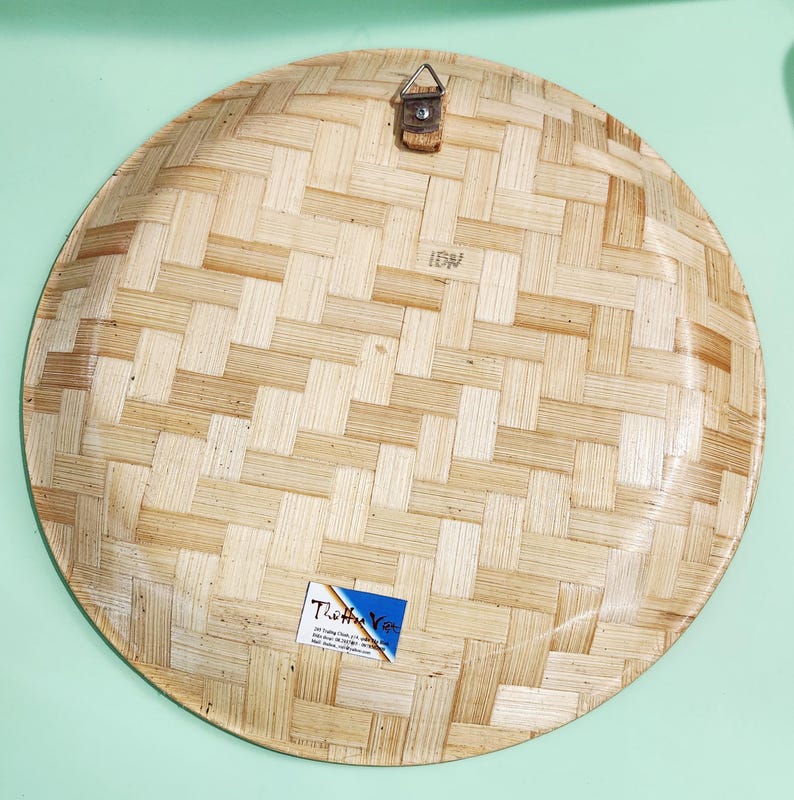 May include: A round, woven bamboo plate with a natural light brown color. The plate has a basket weave pattern and a small metal hanger attached to the top. A small sticker is affixed to the bottom.