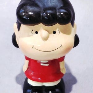 May include: A ceramic figurine of a cartoon character with black hair, a red shirt, and black shoes. The figurine has a smiling face and is standing upright. The overall design is simple and colorful.