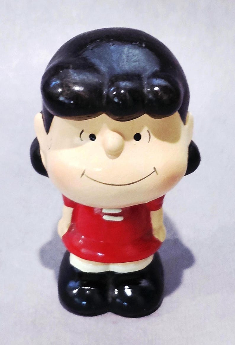 May include: A ceramic figurine of a cartoon character with black hair, a red shirt, and black shoes. The figurine has a smiling face and is standing upright. The overall design is simple and colorful.