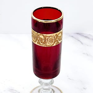 May include: A tall, ruby red champagne flute with a gold rim and a decorative gold band featuring a floral design. The stem is clear glass with a gold accent at the base of the bowl. The glass is elegant and ornate.
