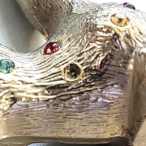 May include: Close-up of a gold-toned, textured metal object with embedded gemstones. The gemstones are various colors, including red, green, and yellow. The metal has a wood-grain appearance.