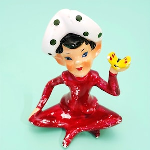 May include: A vintage ceramic figurine of a smiling character in a red dress and a white hat with green polka dots. The figurine is holding a yellow butterfly with red spots. The figure has blue eyes and black hair.