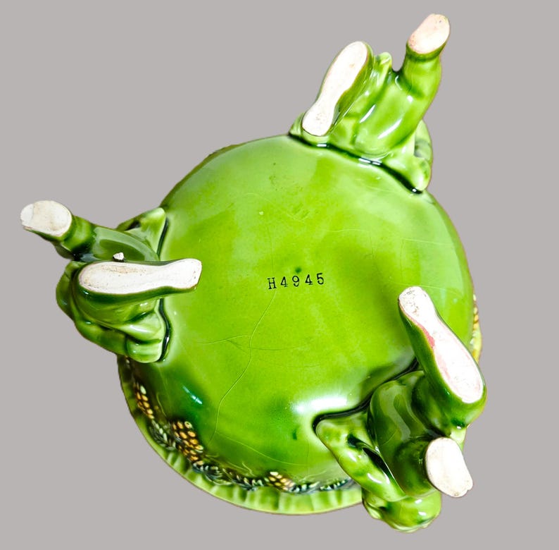 May include: A bright green ceramic bowl viewed from the underside, featuring three sculpted feet and the black printed text H4945. The glossy surface displays a fine network of cracks throughout the glaze, with textured details visible along the edge.