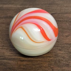 May include: A spherical glass marble featuring a smooth, opaque white base with swirling, translucent ribbons of bright orange and soft pink colors that wrap around the surface in a continuous, flowing pattern, creating a glossy, reflective finish.