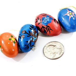 May include: Four hand-painted wooden Easter eggs in orange, red, and blue, each with floral designs. The eggs are strung with gold thread for hanging. A U.S. quarter is included for size reference.