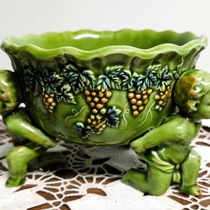May include: A green ceramic bowl featuring a scalloped rim and a relief pattern of grape clusters and leaves. The bowl is supported by two sculpted figures of kneeling cherubs on opposite sides, finished in a matching glossy green glaze.