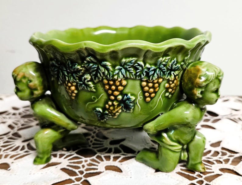 May include: A green ceramic bowl featuring a scalloped rim and a relief pattern of grape clusters and leaves. The bowl is supported by two sculpted figures of kneeling cherubs on opposite sides, finished in a matching glossy green glaze.