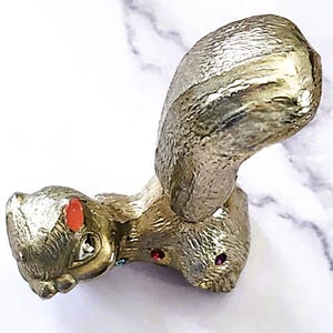May include: A gold-colored squirrel figurine with a textured finish. The squirrel has a large, curved tail and is adorned with red and purple jewel-like accents on its face. The figurine is set against a white marble background.