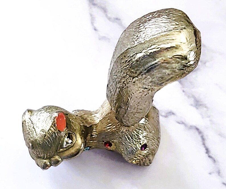 May include: A gold-colored squirrel figurine with a textured finish. The squirrel has a large, curved tail and is adorned with red and purple jewel-like accents on its face. The figurine is set against a white marble background.