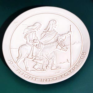 May include: A white ceramic plate with a relief depicting the Flight into Egypt. The image shows two figures, one riding a donkey, and the other walking alongside. The plate has the text "19 - FLIGHT INTO EGYPT - G8" around the edge.