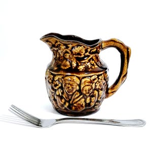 May include: A brown ceramic pitcher with a detailed relief design of figures and foliage. The pitcher has a curved handle and a spout. A silver fork is in the foreground.