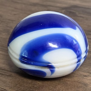 May include: A small, rounded, spherical object made of smooth, polished glass. The surface features a swirling, marbled pattern of opaque white and deep royal blue colors. A horizontal seam runs around the center of the object, indicating two halves.