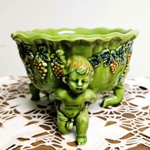 May include: A glossy, light green ceramic bowl featuring a scalloped rim and a relief design of grape clusters and leaves. The bowl is supported by three sculpted cherub figures that serve as feet, each with a smooth, glazed finish.