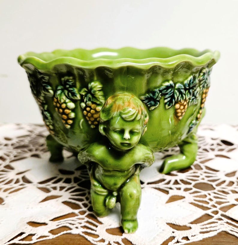 May include: A glossy, light green ceramic bowl featuring a scalloped rim and a relief design of grape clusters and leaves. The bowl is supported by three sculpted cherub figures that serve as feet, each with a smooth, glazed finish.