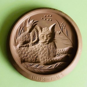 May include: A round, brown ceramic plaque featuring a relief carving of a cat lying down with a small goat positioned behind it. The design includes stylized foliage and the embossed text 1982-1983 along the bottom edge of the circular frame.