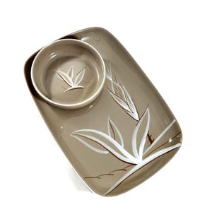 May include: A two-piece serving set featuring a rectangular tray and a round bowl. The set is a muted taupe color with white and brown stylized floral designs. The bowl sits on the tray, and both pieces have a glossy finish.