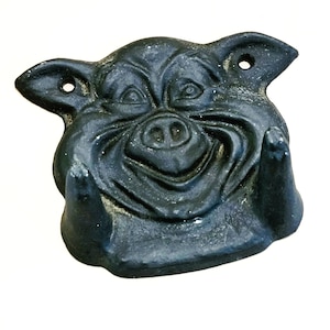 May include: A black, cast iron pig head sculpture with a smiling expression. The sculpture has two holes in the ears for mounting. The pig has visible tusks and wrinkles, giving it a rustic appearance. The sculpture is likely a decorative item.