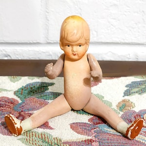 May include: A vintage doll with a painted face, blonde hair, and articulated limbs. The doll is nude, with painted details on the face and limbs. It is wearing white socks and brown shoes. The doll is sitting with its legs spread.