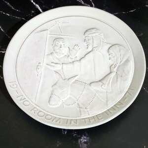 May include: A white ceramic plate with a raised relief scene depicting a biblical narrative. The plate features figures, a donkey, and text that reads "19-NO ROOM IN THE INN-71". The design is monochromatic.