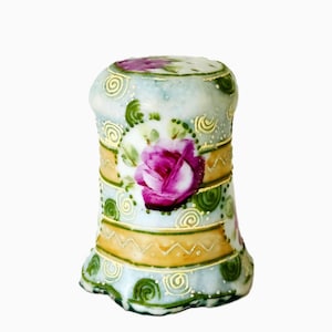 May include: A vintage porcelain thimble with a hand-painted floral design. The thimble features a light blue base with a pink rose, gold accents, and green swirls. The thimble is a collectible item.