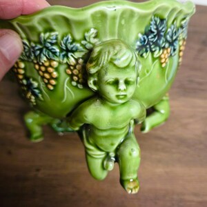 May include: A glossy green ceramic bowl featuring a relief sculpture of a cherub figure on the exterior. The bowl is decorated with raised grape clusters and vine leaf patterns around the rim, supported by three cherub-shaped feet.