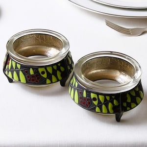 May include: Two circular glass bowls with metallic rims, featuring a black exterior decorated with a repeating pattern of bright lime green leaf shapes and clusters of small dark red berries, each supported by three short, sturdy black feet.