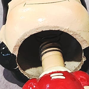 May include: A ceramic figurine with a spring-loaded neck. The figurine's head is cream-colored with black accents, and it wears a red outfit with white stripes. The interior of the figurine is dark, revealing a metal spring mechanism.