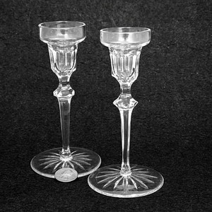 May include: Two clear glass candle holders with a starburst design on the base. The candle holders have a slender stem and a small, round cup to hold a candle. The glass is clear and reflects light.