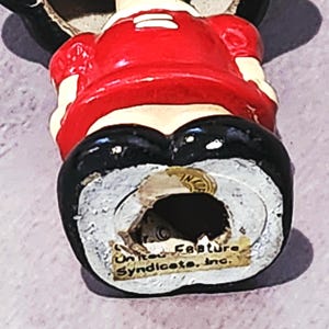 May include: A vintage figurine of a cartoon character, wearing a red top, black shorts, and black shoes. The figurine has a white face and is marked "United Feature Syndicate, Inc."
