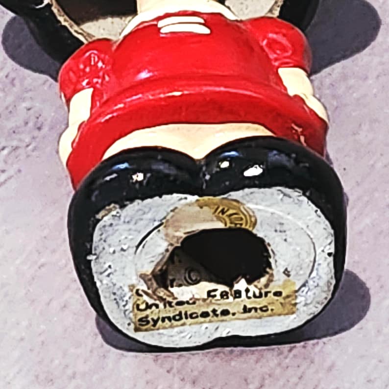 May include: A vintage figurine of a cartoon character, wearing a red top, black shorts, and black shoes. The figurine has a white face and is marked "United Feature Syndicate, Inc."