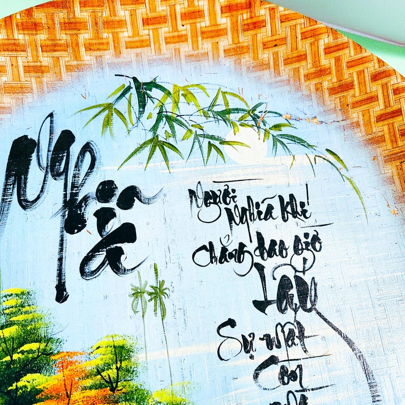 May include: Round, hand-painted artwork with a bamboo and landscape design. The artwork features calligraphy in black ink on a light blue background. The outer edge is woven with a natural, light brown material.