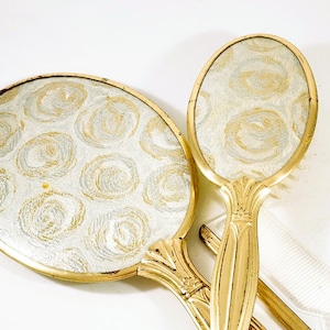 May include: A vintage vanity set featuring a hand mirror and a brush. The set has gold-toned frames and handles. The mirror and brush backs have a white and gold floral pattern. The brush has white bristles and a white comb is visible.