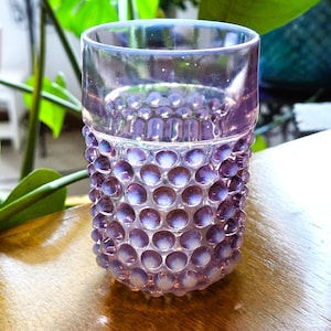 May include: A translucent lavender glass tumbler featuring a hobnail texture with raised circular bumps covering the lower two-thirds of the exterior. The upper rim is smooth and clear, while the base has a rounded shape with a consistent purple hue.
