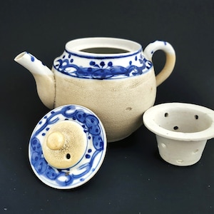 May include: Ceramic teapot with a lid and infuser. The teapot is beige with blue floral designs around the rim and on the lid. The spout and handle are white with blue accents. The infuser is white with several holes.