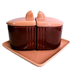 May include: A set of two dark brown ceramic containers with light pink lids, each featuring a small vertical handle. The containers are placed side-by-side on a matching light pink square ceramic tray, creating a cohesive two-piece set.