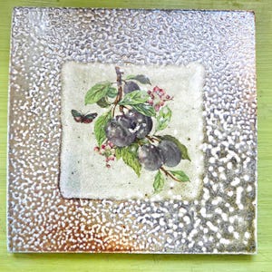 May include: A square decorative tile featuring a central illustration of dark purple plums on a branch with green leaves, small pink flowers, and a tiny butterfly. The border has a textured, mottled metallic finish in shades of silver and bronze.