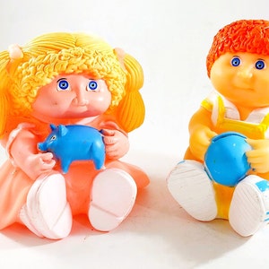 May include: Two vintage Cabbage Patch Kids dolls. The girl doll has blonde hair and is holding a blue piggy bank. The boy doll has red hair and is holding a blue ball. Both dolls are wearing white shoes.