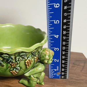 May include: A light green ceramic bowl featuring a scalloped rim and a relief design of grape clusters and leaves. A cherub figure supports the bowl from the side. A blue ruler is positioned next to the bowl, showing a height of approximately 3 inches.