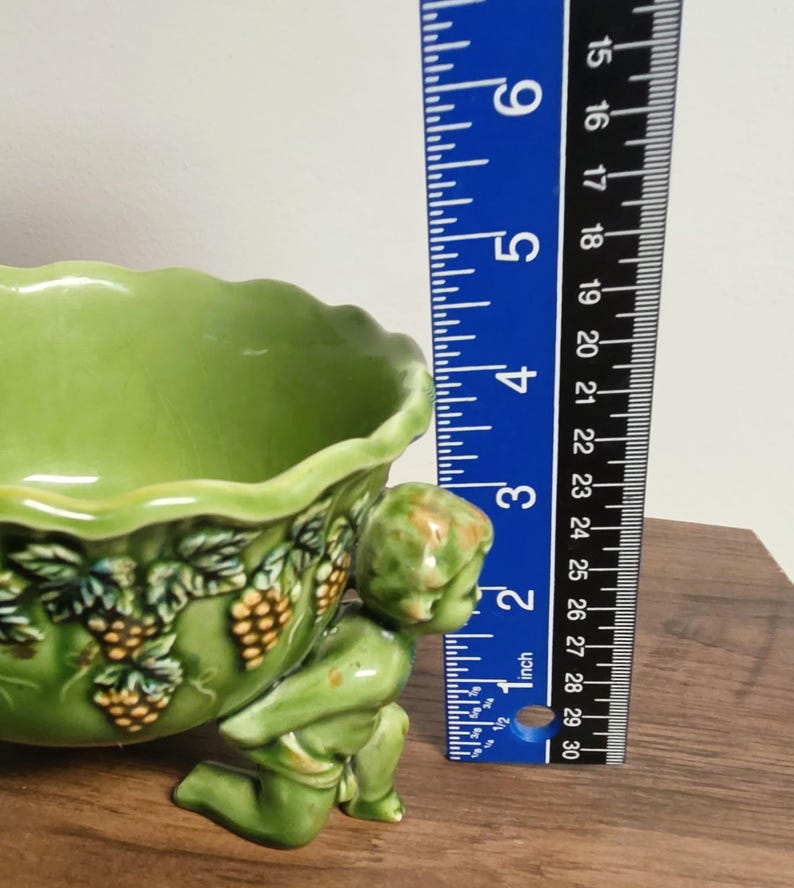 May include: A light green ceramic bowl featuring a scalloped rim and a relief design of grape clusters and leaves. A cherub figure supports the bowl from the side. A blue ruler is positioned next to the bowl, showing a height of approximately 3 inches.