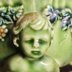 May include: A glossy green ceramic figurine featuring a sculpted cherub-like face with soft facial features. The background displays a raised relief pattern of green leaves and small clusters of light-colored grapes, all finished with a smooth, reflective glaze.