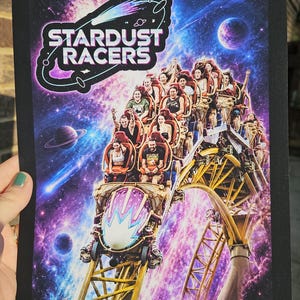Könnte beinhalten: Ein rechteckiger Druck mit dem Text STARDUST RACERS in weißen Buchstaben vor einem violetten und blauen kosmischen Hintergrund. Das Bild zeigt einen Achterbahnzug voller Fahrgäste, der auf gelben Schienen durch das Weltall fährt.