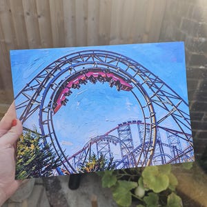 May include: A rectangular print featuring an oil painting style depiction of a red roller coaster train navigating a large circular loop. The metal track structure is dark gray against a bright blue sky, with green foliage visible at the bottom.